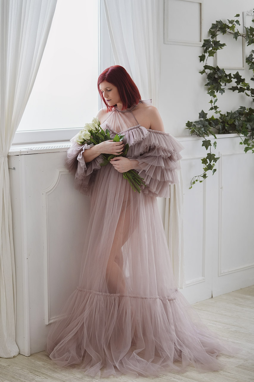 Ablak mellett álló lila ruhás nő rózsacsokrot tart a karján