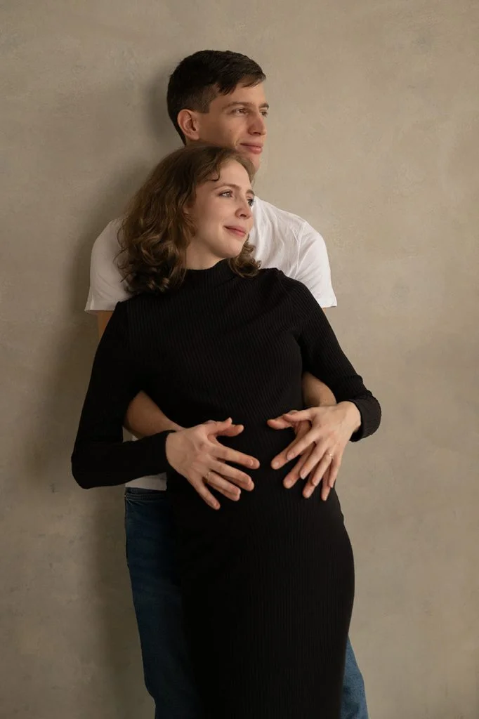 A pár egymáshoz simulva áll egy elegáns, bézs fal előtt, a kismama fekete ruhát visel.
