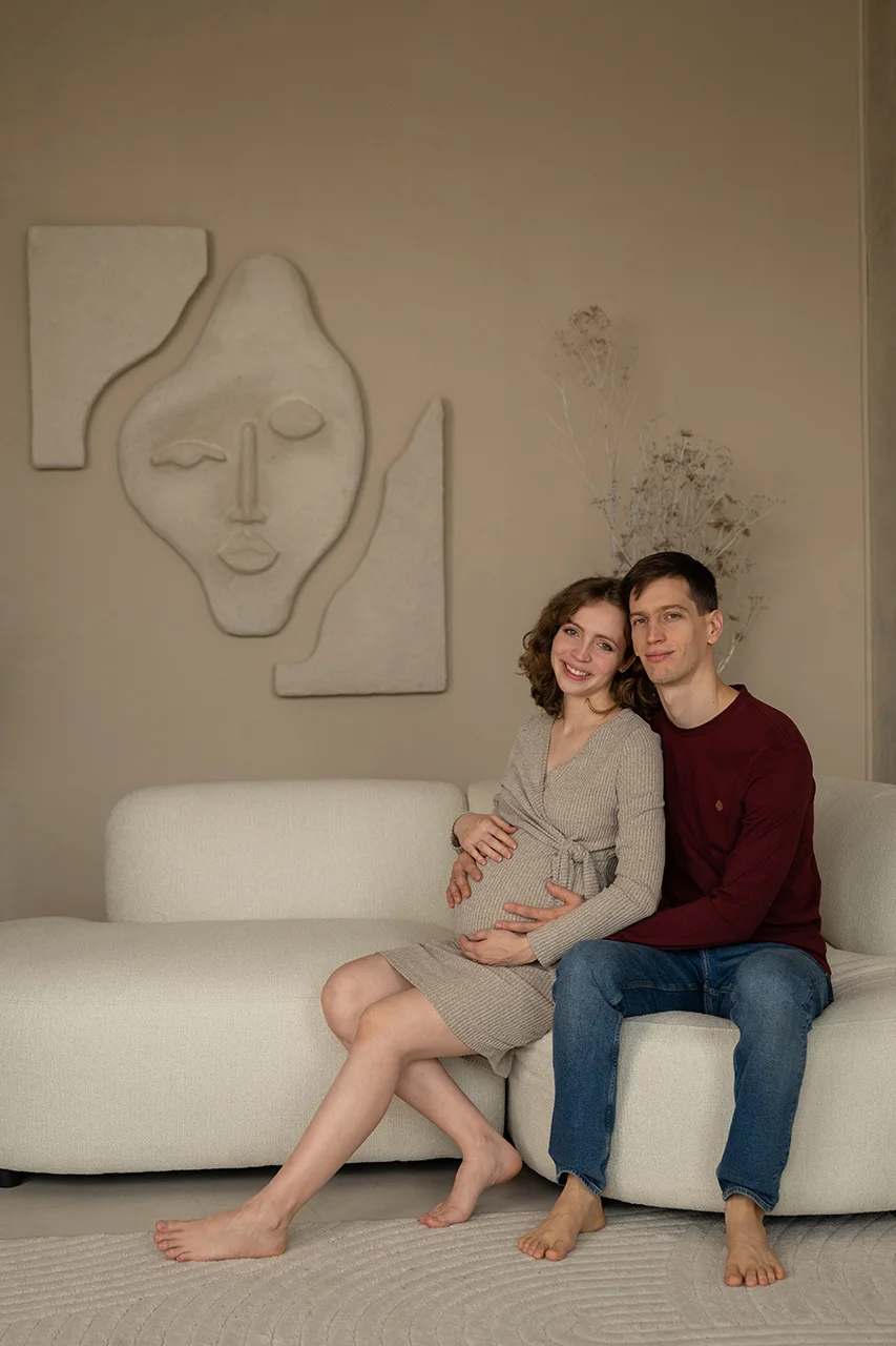 Pár kanapén ülve, egymást átölelve, kismama pocakját simogatva.