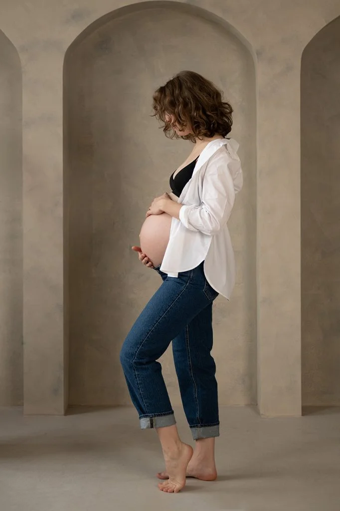 Kismama fekete melltartóban és fehér ingben, farmerben, oldalt állva, kezét a pocakján tartva.