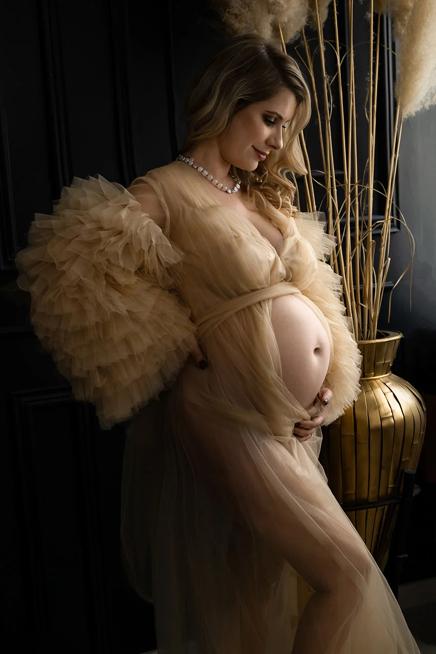 Kismama bézs tüllruhában, megvilágított pocakkal, elegáns stúdió környezetben.