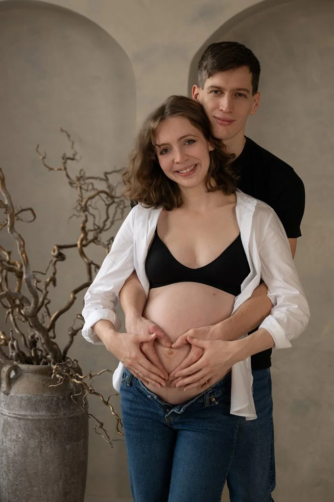 Pár keze szív alakban a várandós pocakon, mosolygó kismamával előtérben.
