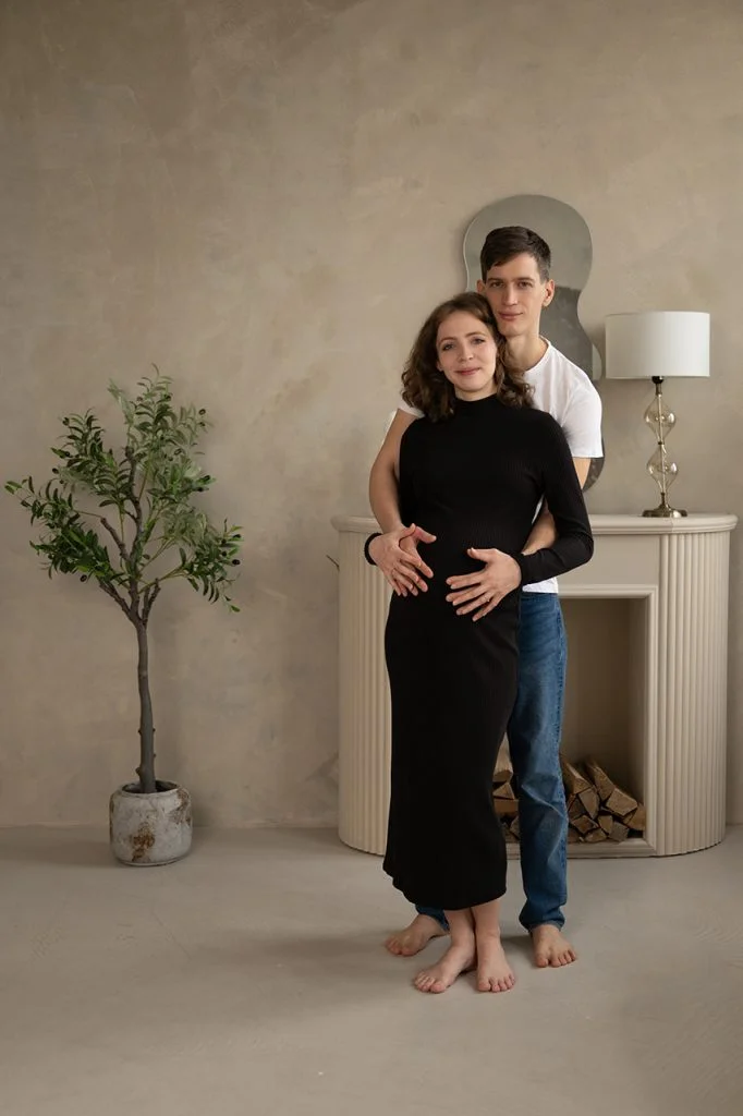 A pár egymáshoz simulva áll egy elegáns, bézs fal előtt, a kismama fekete ruhát visel.